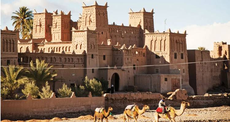 Mud-brick towers of the fortified Ksar of Aït Benhaddou stand under a blue sky while camel riders pass in the foreground.