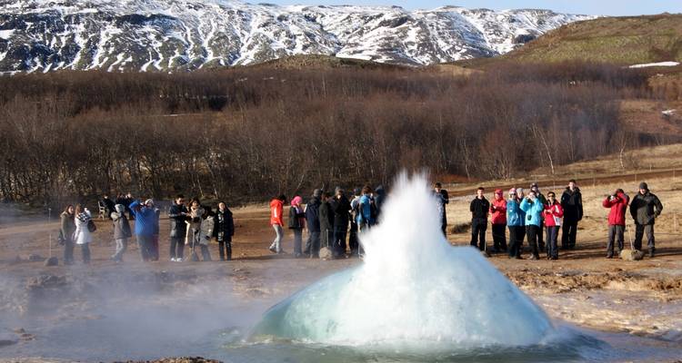 People gathered around a geyser set against a backdrop of mountains