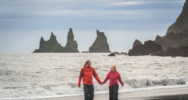 Pareja caminando por una playa de arena negra con farallones al fondo.