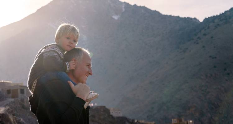 Een ouder die een kind op de schouders draagt met bergen op de achtergrond.