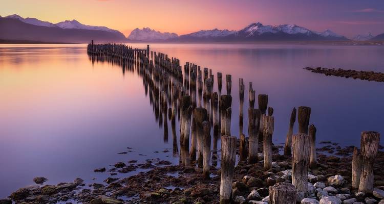 Serene view of old wooden posts in a calm body of water with distant snow-capped mountains at sunset.
