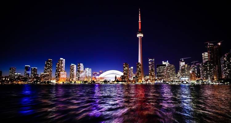 Nighttime city skyline with skyscrapers and CN Tower reflecting on water.