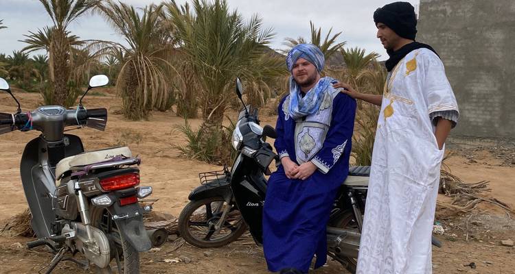 Two men in traditional clothing sitting on motorcycles in a desert environment.
