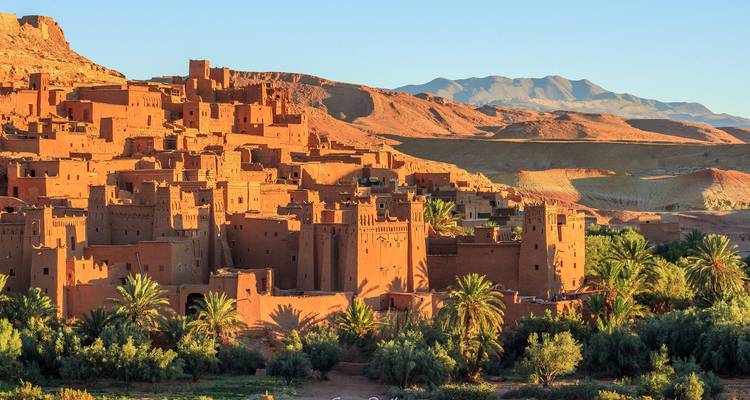 Pueblo fortificado de adobe y palmeras al atardecer con montañas desérticas distantes.
