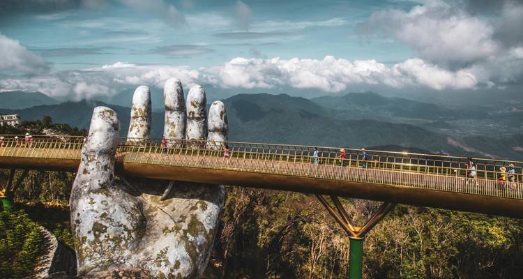 Gouden brug vastgehouden door reuzenachtige handen met bergachtige achtergrond onder bewolkte hemel.