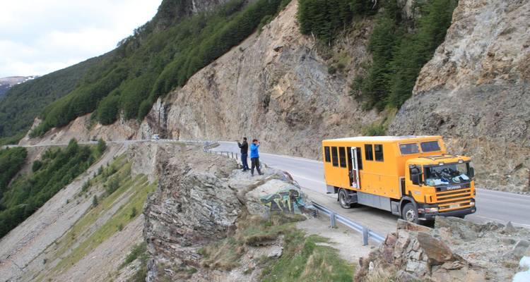 An overland truck parked on a cliffside with two people taking photos.
