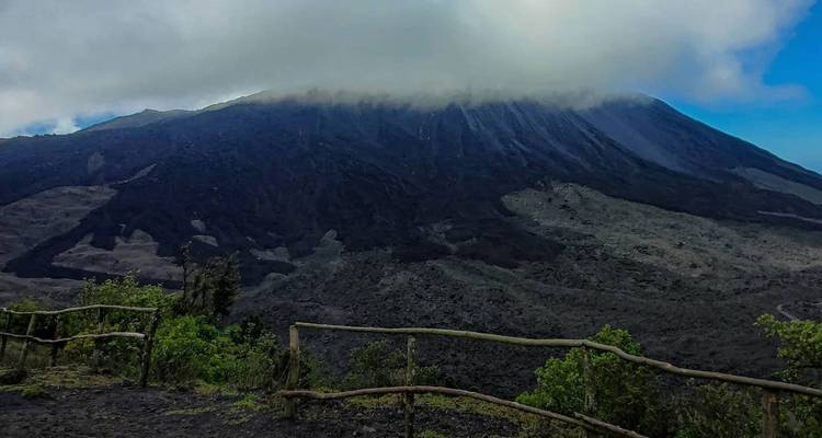 Pico volcánico coronado de nubes con campos de lava árida vistos desde un mirador con cerca de madera rústica.