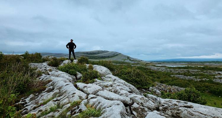 Randonneur en solo debout sur un pavage calcaire dominant un paysage verdoyant vallonné dans le Burren sous un ciel couvert
