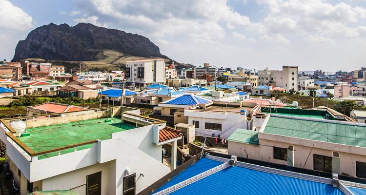 Techos coloridos del pueblo costero de Jeju bajo un dramático cono de toba volcánica y nubes dispersas