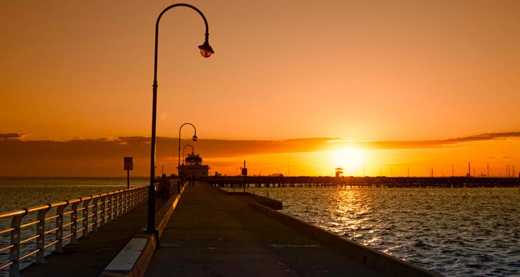 Long pier avec des lampadaires vintage s'étendant dans les eaux océaniques calmes lors d'un coucher de soleil d'un orange éclatant.