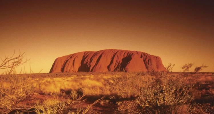 Monolithe de grès rouge massif Uluru s'élevant des plaines désertiques plates sous une lumière chaude du soir.