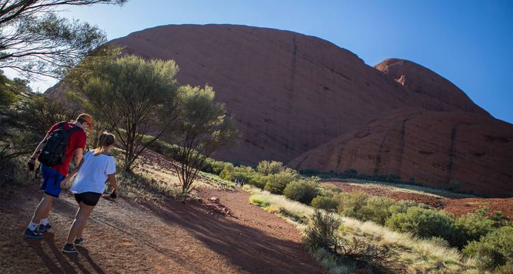 Deux randonneurs marchent le long d'un sentier poussiéreux bordé de majestueuses formations rocheuses rouges et d'arbres eucalyptus clairsemés.