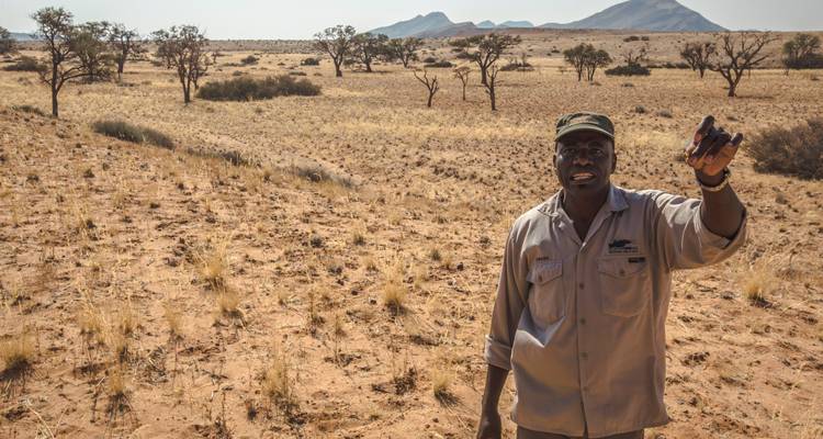 Safarigids staat in halfwoestijnachtige savanne en houdt een voorwerp omhoog terwijl hij het landschap uitlegt.