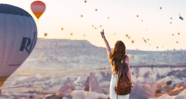 Mujer joven con mochila señalando hacia docenas de globos aerostáticos flotando sobre Capadocia.
