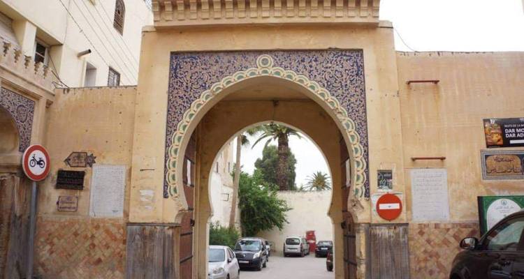 Une arcade carrelée décorative mène à un parking de cour avec un palmier au-delà.