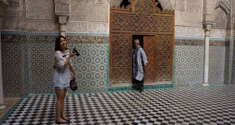 Un touriste tient un appareil photo dans la cour carrelée luxueusement d'une médersa tandis qu'un ancien en djellaba observe depuis un portail sculpté.