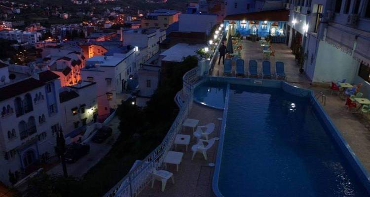 Vue nocturne des lumières d'une ville perchée se reflétant près d'une piscine sur toit bordée de chaises.