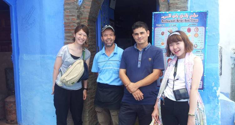 Quatre voyageurs sourient devant une porte bleu vif de Chefchaouen à côté d'un panneau de menu touristique.