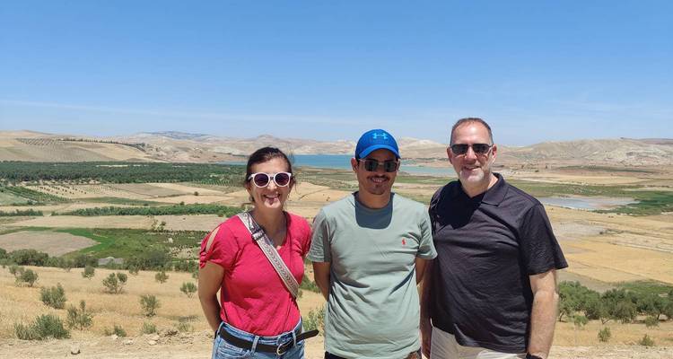 Trois voyageurs se tiennent souriants devant un paysage doré vallonné et un réservoir au loin sous un ciel bleu dégagé.