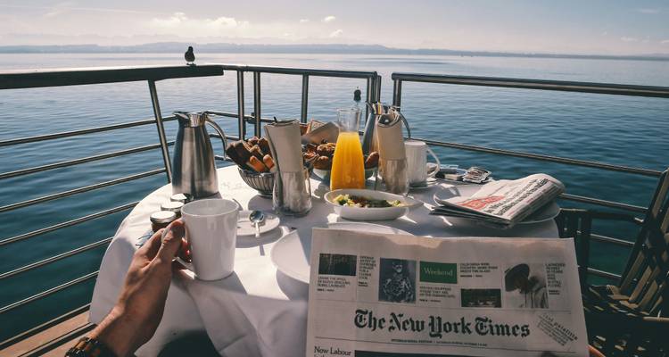 Mesa de desayuno en una terraza con vista a un mar tranquilo con una persona leyendo un periódico.