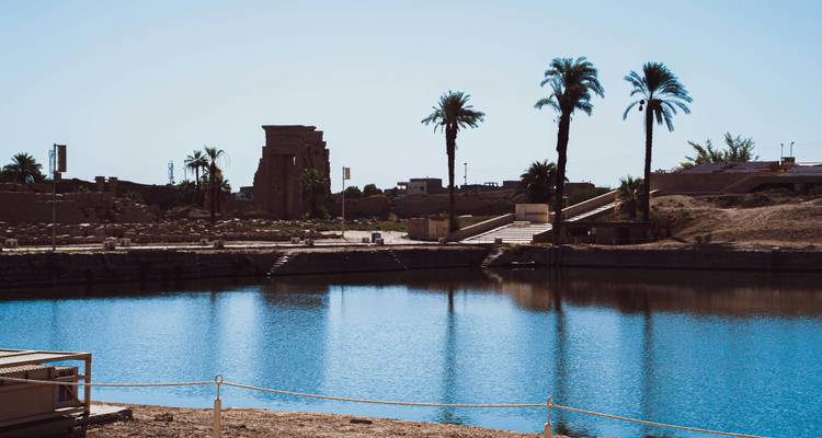 Ruinas de un templo y una piscina reflectante con palmeras y cielo azul.