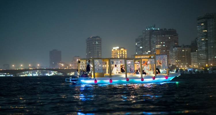 Un barco iluminado en un río por la noche con el horizonte de la ciudad al fondo.