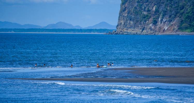 Een schilderachtig uitzicht op een strand met mensen in het water en weelderige groene kliffen.