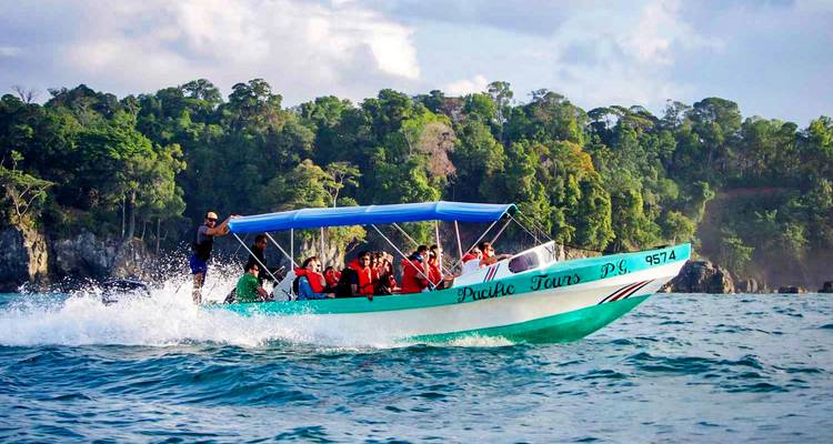 Barco turístico con personas navegando a alta velocidad cerca de una costa boscosa.