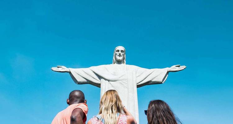 Reisende blicken zu der hoch aufragenden Christus-Erlöser-Statue gegen einen klaren blauen Himmel empor.