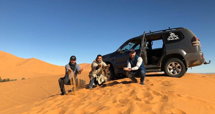 Trois hommes jouant avec le sable sur une dune avec un véhicule Dakar.