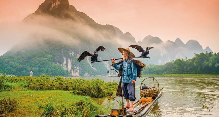 Man fishing on a wooden raft with cormorants, misty mountains in the background