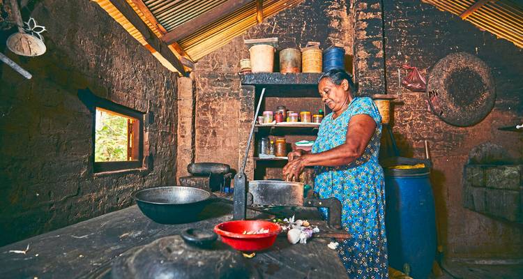 A Sri Lankan woman prepares traditional dishes at a rustic clay-brick kitchen table lined with spices.