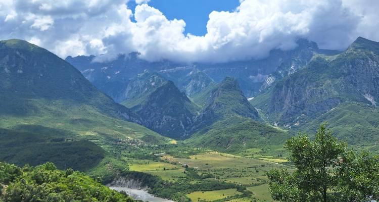 Valle albanés que se extiende ampliamente con un río sinuoso flanqueado por montañas verdes dramáticas y picos coronados de nubes.