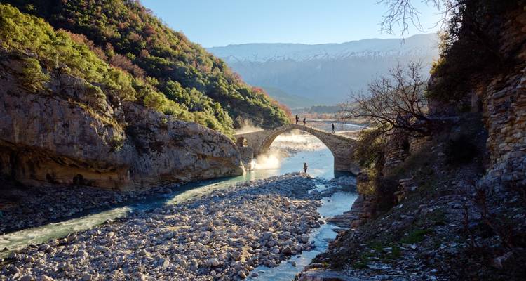 Puente de arco de piedra histórico que atraviesa un río turquesa en un cañón estrecho, con gente disfrutando de la vista.