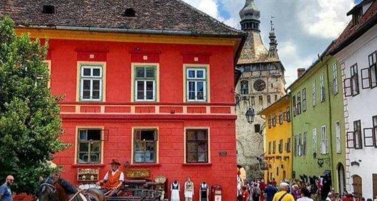 Plaza medieval colorida en Sighisoara con casas vibrantes rojas, amarillas y verdes, carruaje de caballos y turistas paseando.