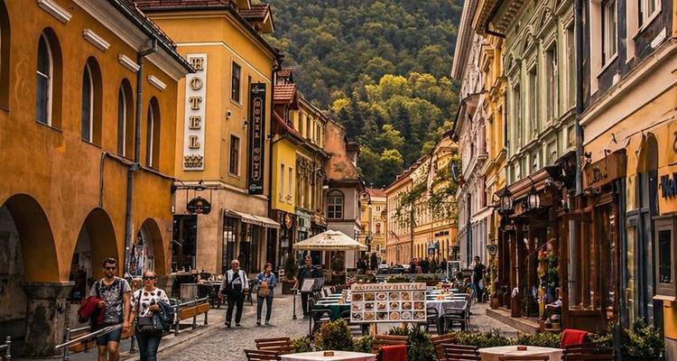 Calle empedrada concurrida en Brasov bordeada de edificios históricos coloridos, cafés al aire libre y colinas boscosas en el fondo.