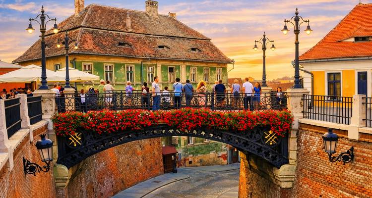 Puente de hierro fundido cargado de flores en Sibiu abarrotado de gente al atardecer, cielo cálido brillando detrás de antiguos tejados.