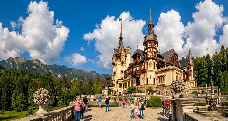 Le château de Contes de fées de Peles entouré par les montagnes boisées des Carpates sous un ciel bleu éclatant, tandis que les touristes explorent la terrasse.