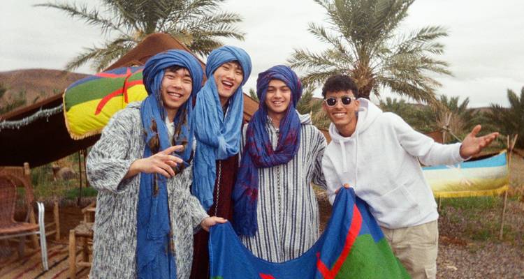 Vier jongeren die traditionele kleding en hoofddoeken dragen staan samen buiten met palmbomen op de achtergrond.