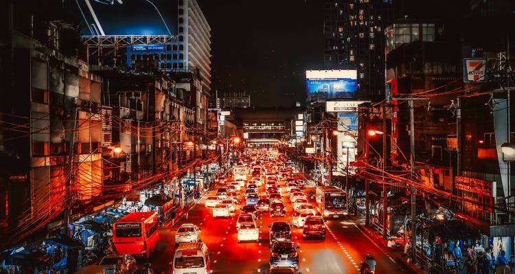 Nächtliche Stadtlandschaft mit starkem Verkehr auf einer belebten Bangkoker Straße, beleuchtet durch rote Rücklichter und Neonlichter.