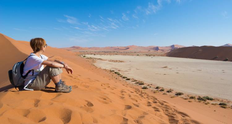 Le voyageur est assis sur la pente d'une dune orange surplombant un vaste playa d'argile dans le désert du Namib sous un ciel dégagé.