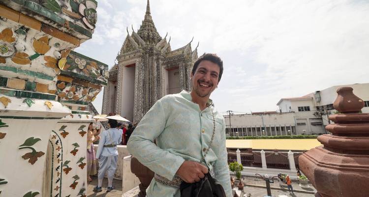 Viajero sonriente con camisa tradicional de pie en los escalones del templo Wat Arun con una aguja ornamentada detrás.