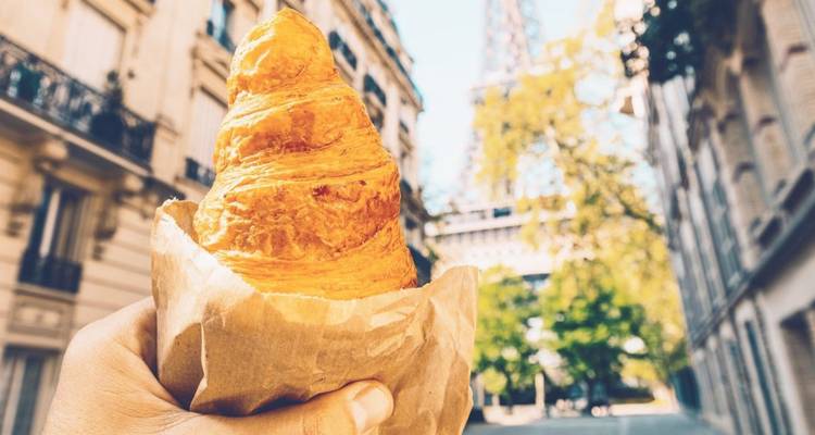 Hand houdt boterige croissant vast met onscherpe Eiffeltoren die tussen Parijse straten uittorent