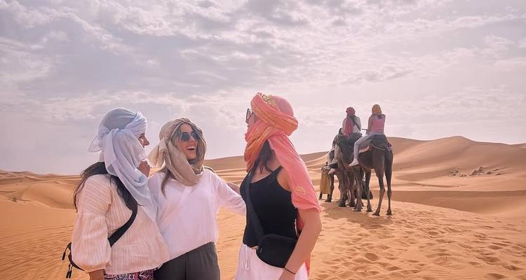 Trois femmes rient ensemble en portant des foulards désertiques tandis que des chaméliers passent derrière elles sur les dunes.