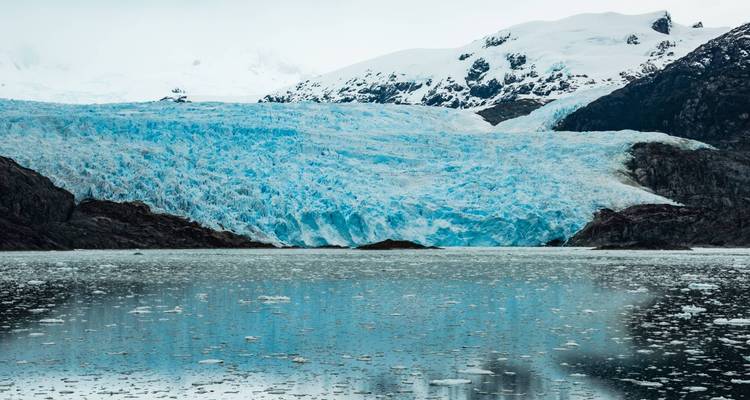 Een gletsjer met ijsblauwe tinten tegen een besneeuwde bergachtergrond.