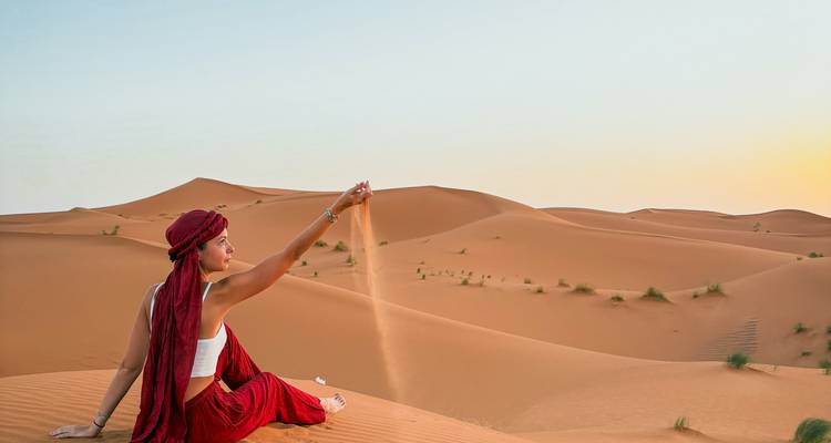 Une femme vêtue de rouge est assise sur une dune au lever du soleil, laissant le sable s'échapper de sa main sur le vaste panorama du désert.