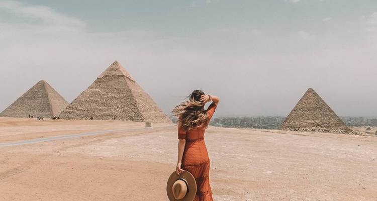 A person in a dress holding a hat, facing the pyramids.