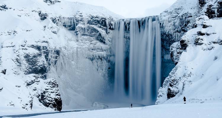 La cascade glacée de Skogafoss en hiver avec de minuscules visiteurs debout sur le sol enneigé de la vallée.