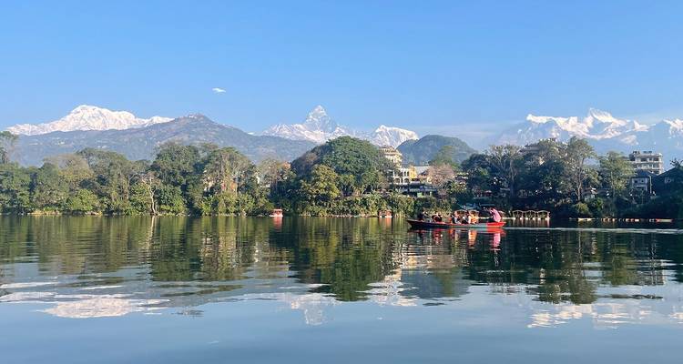 Lago Phewa sereno con botes, orilla boscosa y picos nevados del Himalaya reflejados en el agua