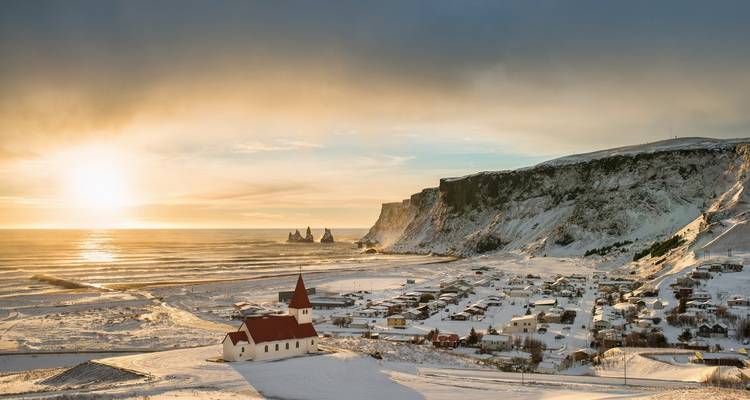 Village enneigé près d'une falaise avec une église au coucher du soleil.